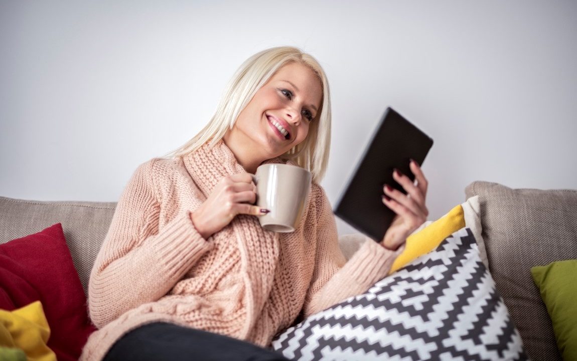 Blonde woman sitting on the sofa inside her new home and holding a cup while watching multimedia content on digital tablet.